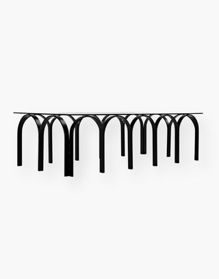 Table with rectangular industrial steel tubing finished in black and is fashioned into a repeating horseshoe arch design and a simple glass top.