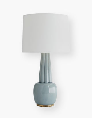 Table lamp with elongated ribbed neck on a rounded base with a gold-finished platform and an off-white linen drum shade.